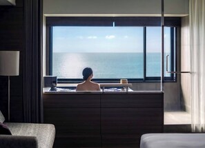 Una persona in vasca da bagno con vista sul mare attraverso un'ampia finestra.
