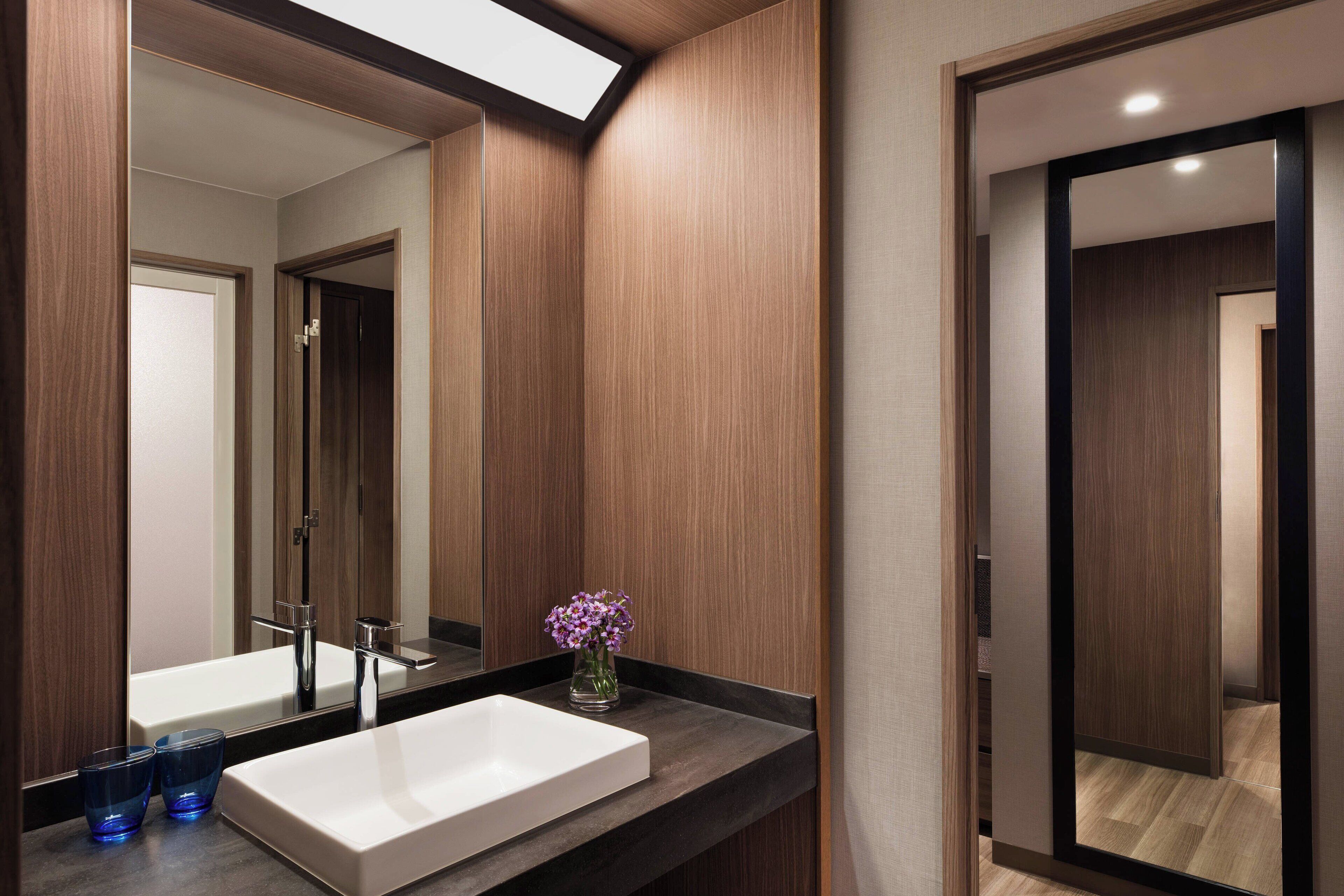 Une salle de bain moderne avec un grand miroir, un lavabo blanc et un vase rempli de fleurs violettes.