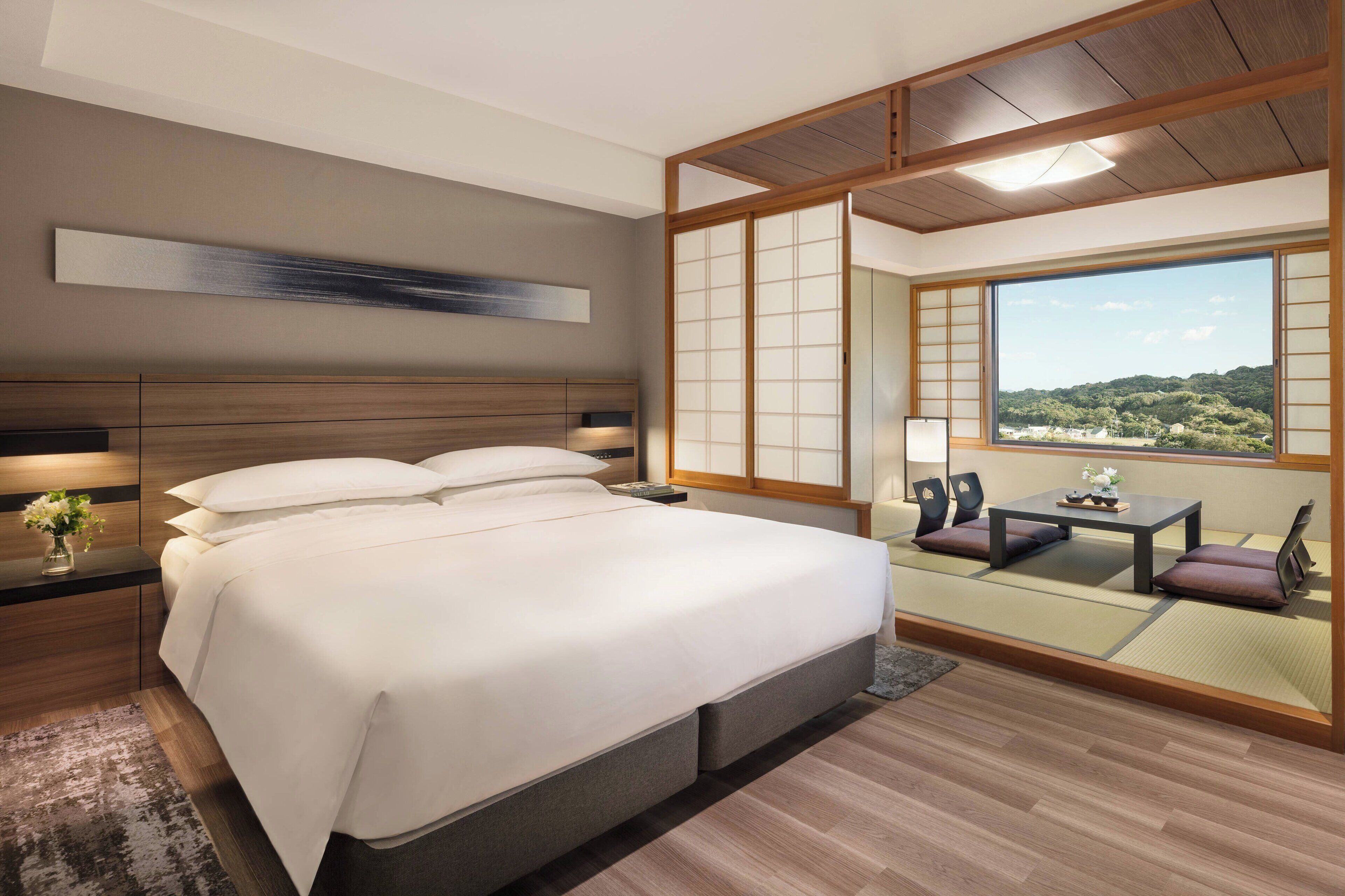 A modern bedroom with a large bed, a nightstand, a bedside table with a vase of flowers, and a view of a scenic landscape through a sliding door.
