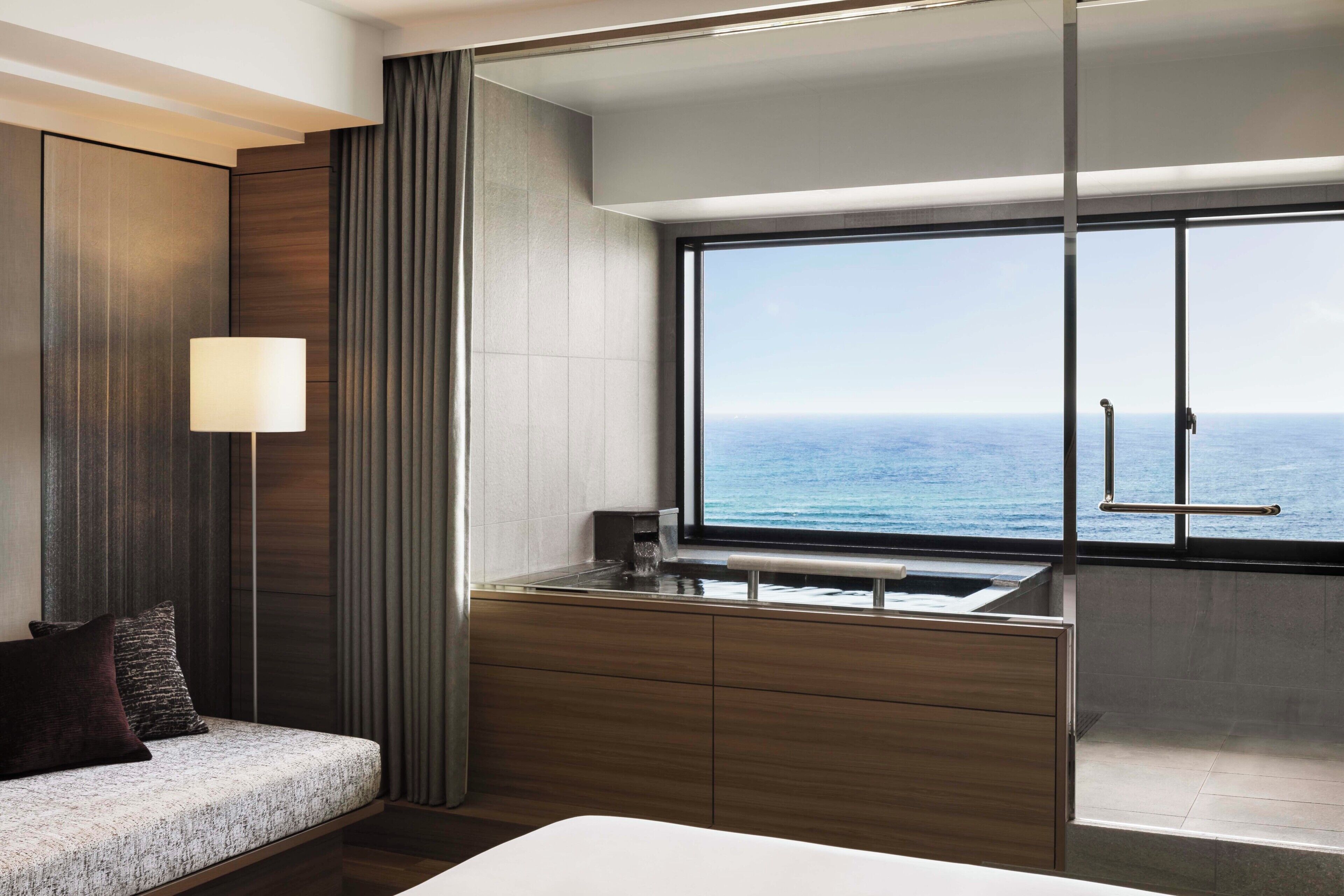 A modern hotel room with a large window, a sofa, a bedside table, and a view of the ocean.