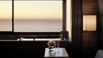 A room with a large window overlooking the sea, a small table with a vase of flowers and two glasses, and a lamp on the side.