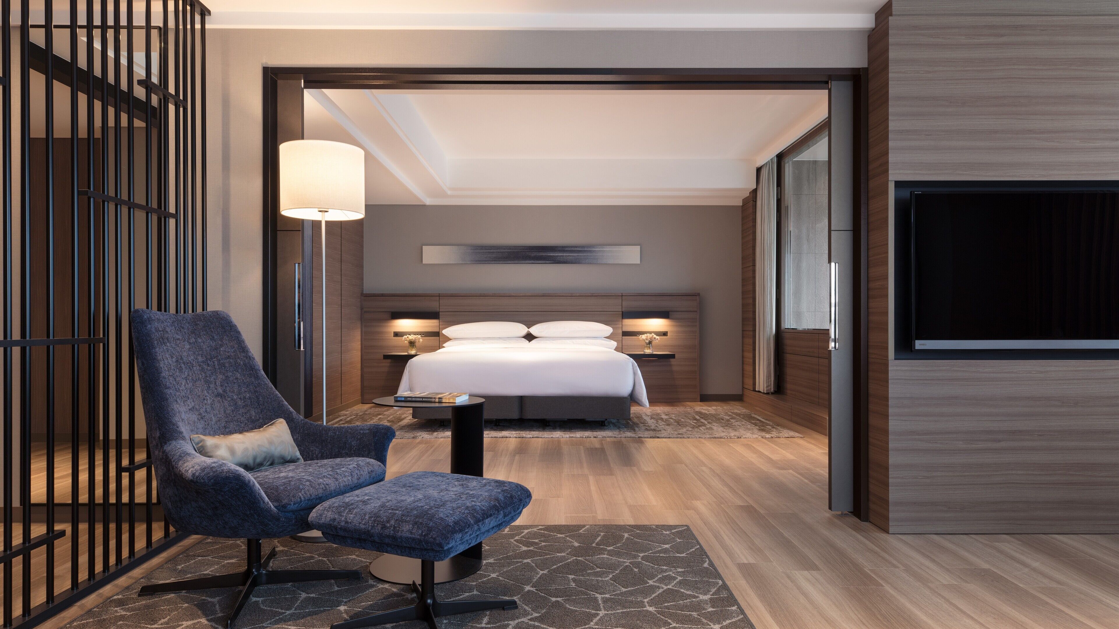 Ein modernes Hotelzimmer mit Bett, einer Sitzecke mit Sessel und Hocker, einem Nachttisch mit Lampe und einem an der Wand montierten Fernseher.