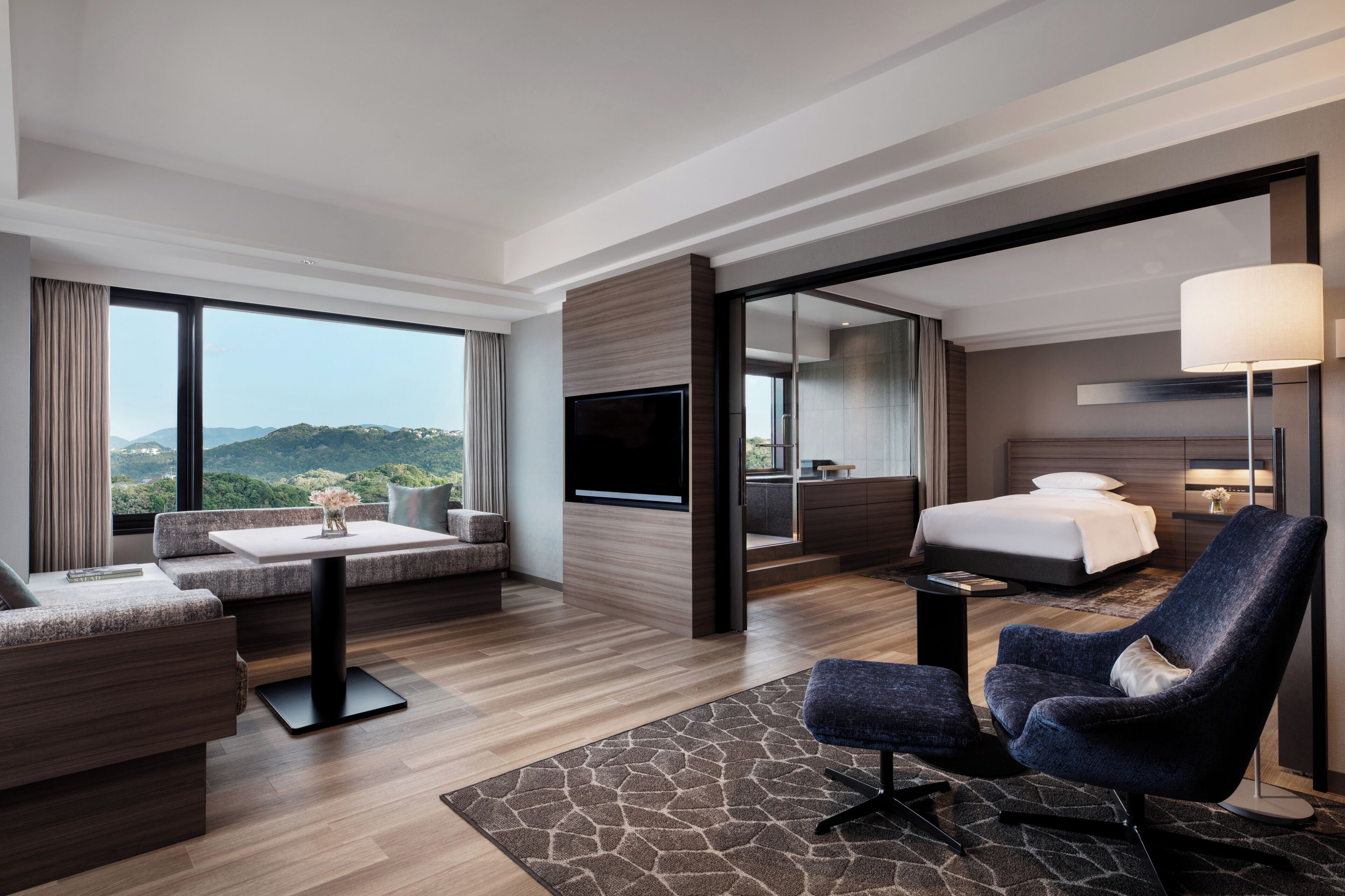 A modern hotel room with a large bed, a sofa, a chair, a flat-screen TV, and a view of the outdoors.