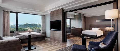 A modern hotel room with a large bed, a sofa, a chair, a flat-screen TV, and a view of the outdoors.