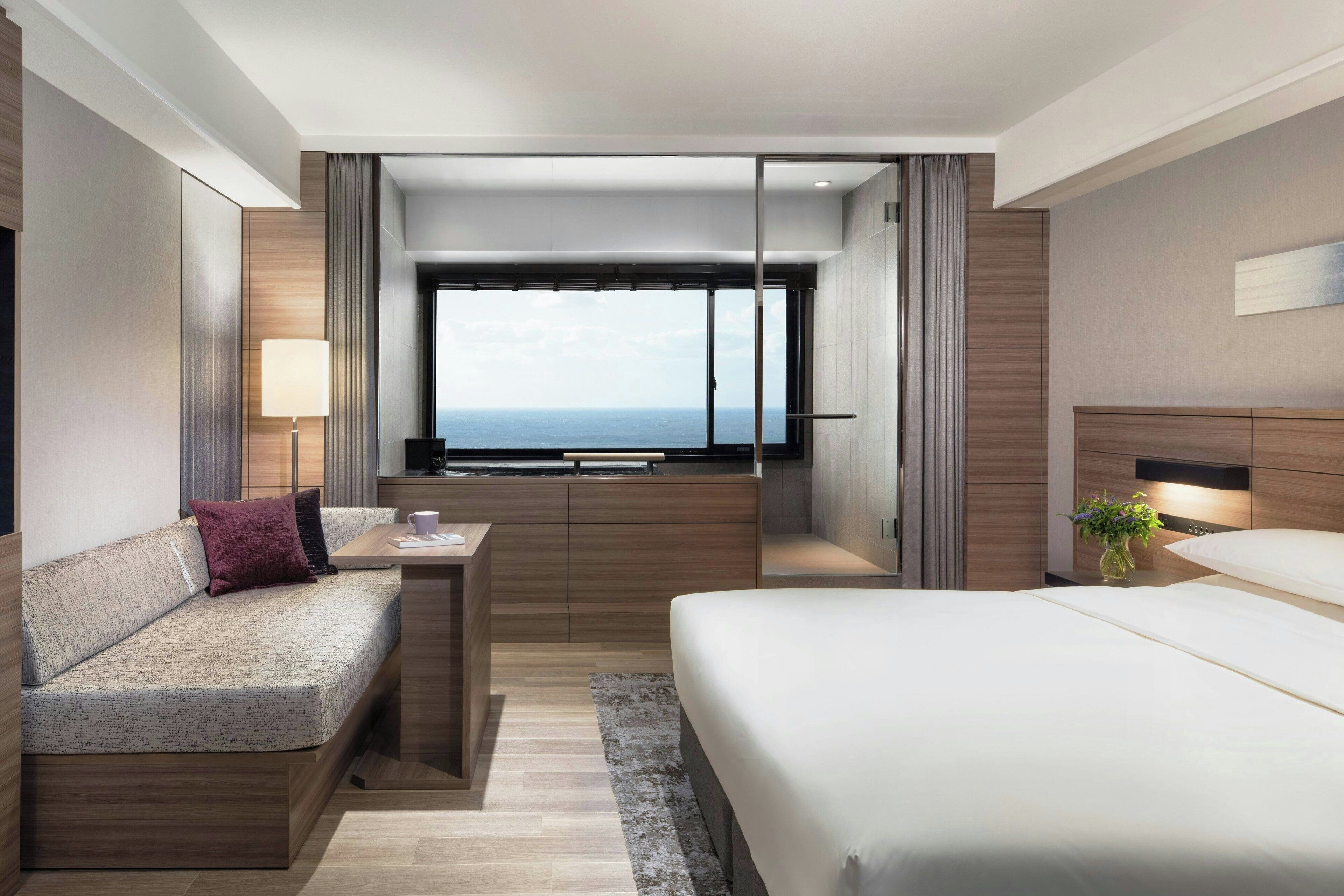 A modern hotel room with a large bed, a sofa, a desk, and a view of the sea.
