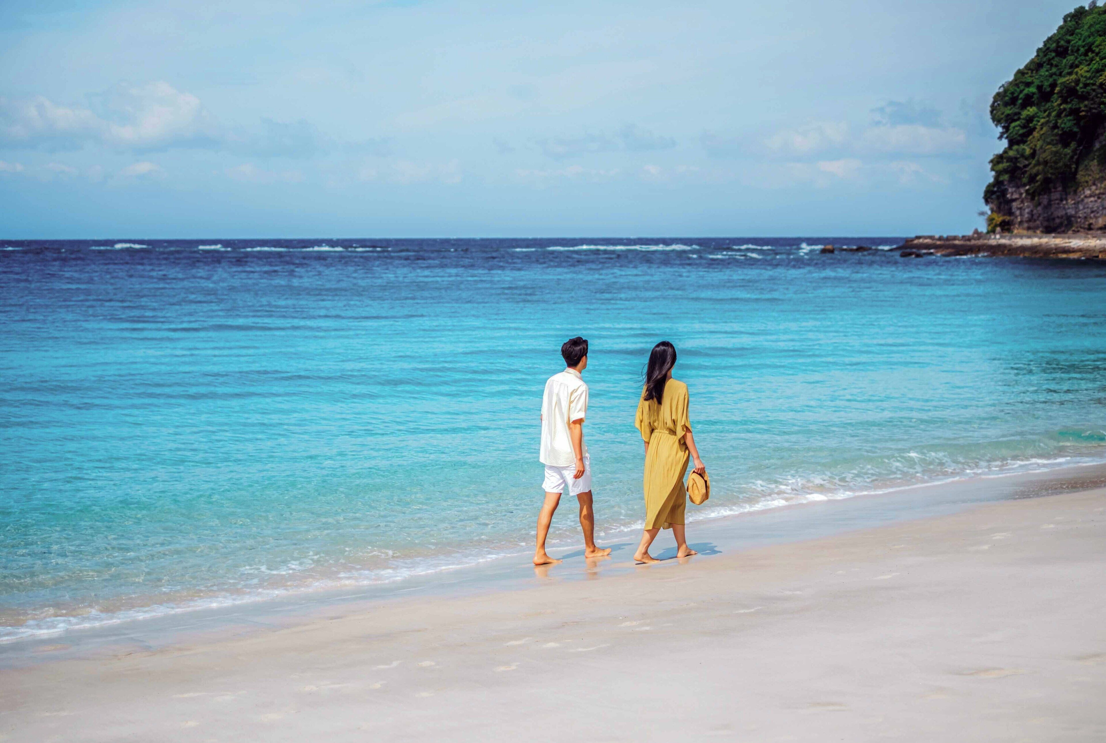 A couple walking along a beach with clear blue water and a rocky cliff in the background.