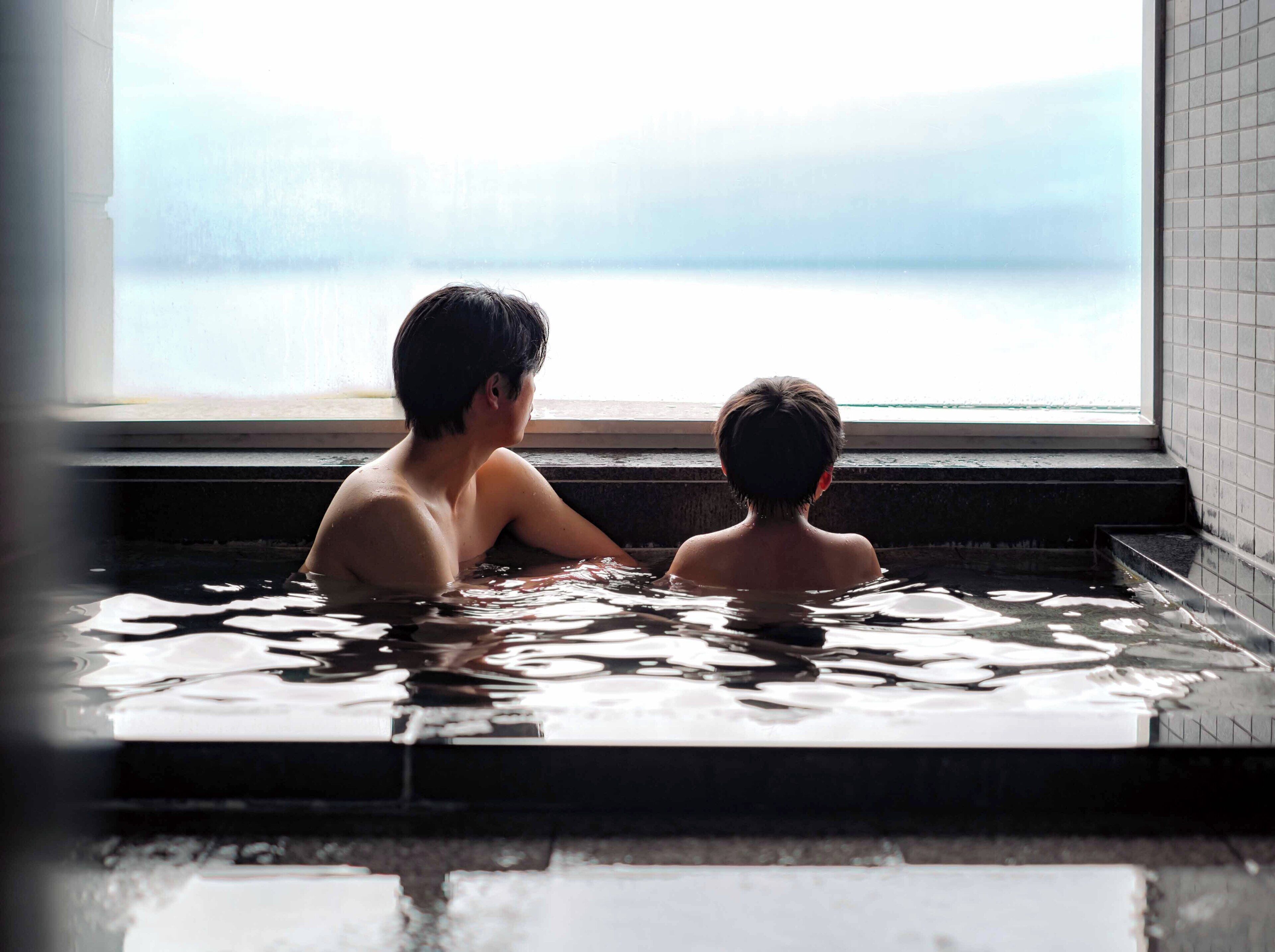 Two individuals in a hot tub with a view of a body of water through a window.
