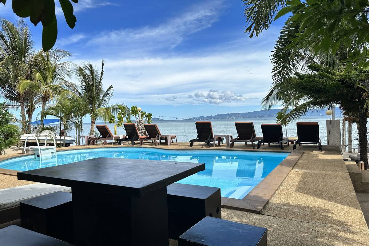 A poolside area with lounge chairs and a table, overlooking a body of water and a distant mountain range.