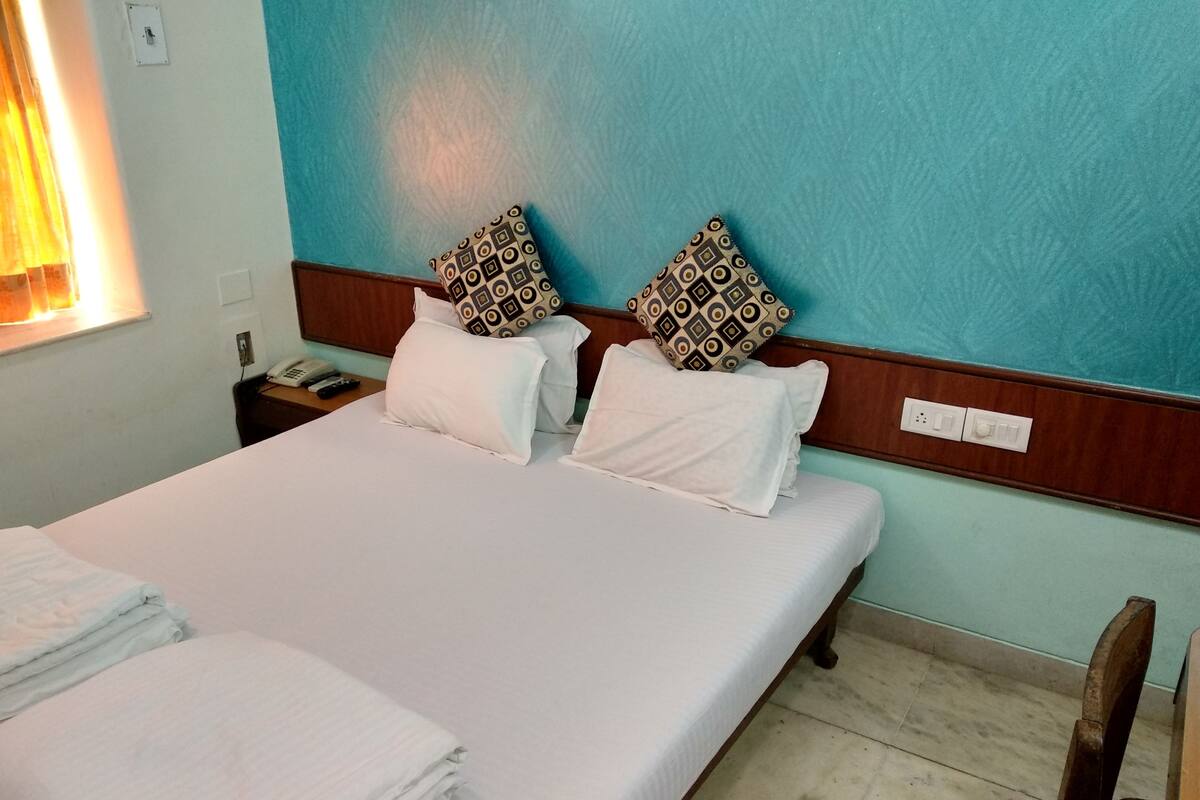 Una cama bien hecha con sábanas blancas y almohadas estampadas, un cabecero de madera, una mesita de noche con teléfono y control remoto, y una ventana con cortinas.