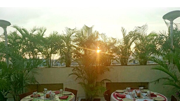 An outdoor dining area with tables set for two, surrounded by potted plants and palm trees. - Hotel Mogul Palace (Mumbai)