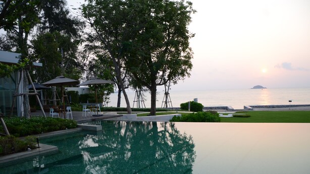 A serene pool area with a view of the sea and a distant island, surrounded by trees and outdoor furniture. - Hua Hin Luxury Beachfront Condo By Mon (Hua Hin)