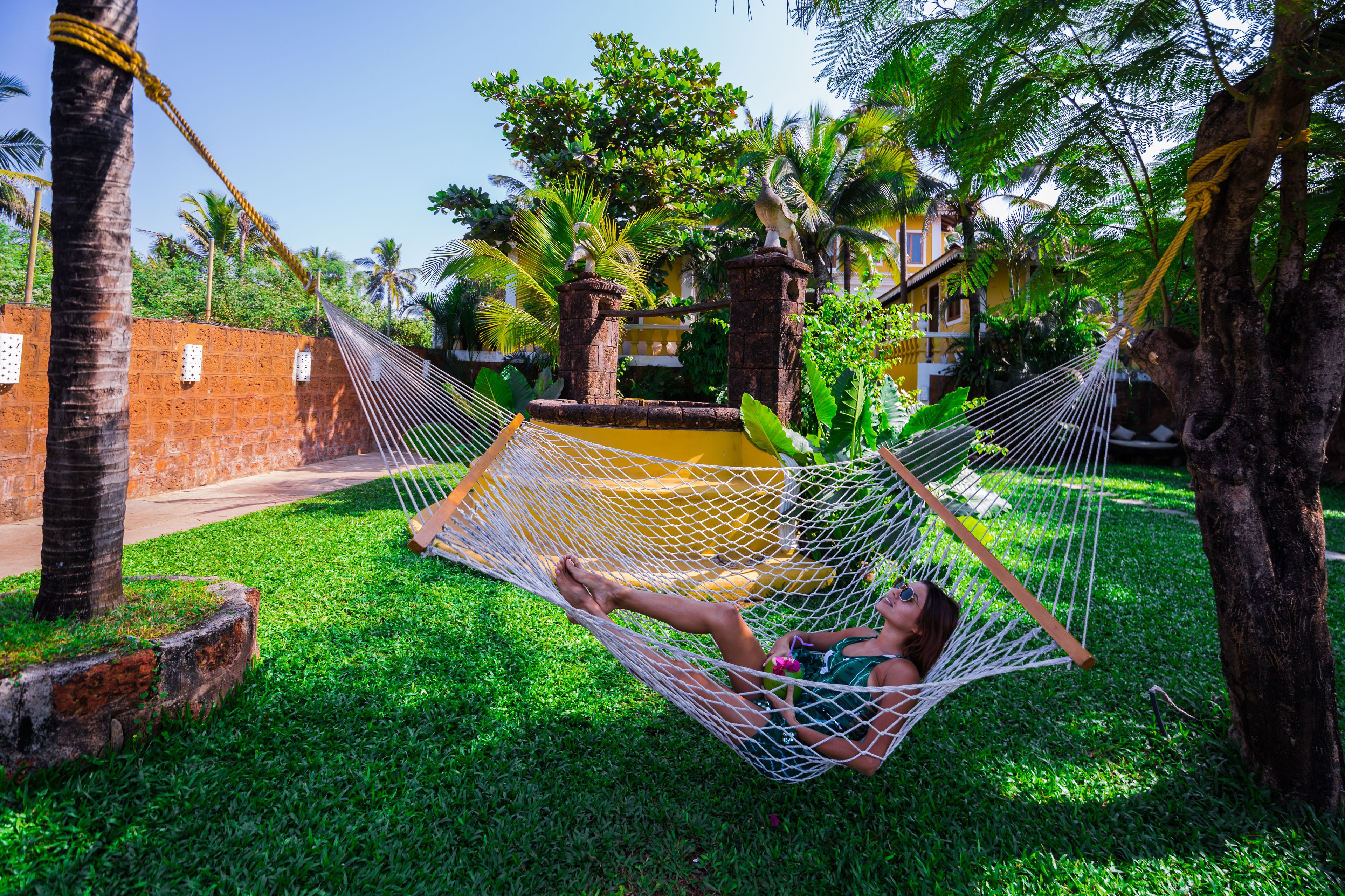 A hammock in a lush garden with palm trees and a brick wall.