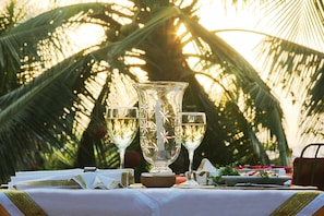 A table set with a large decorative vase, two wine glasses, and a bowl of fruit, with palm trees in the background. - Fiesta Beach Resort (Baga)