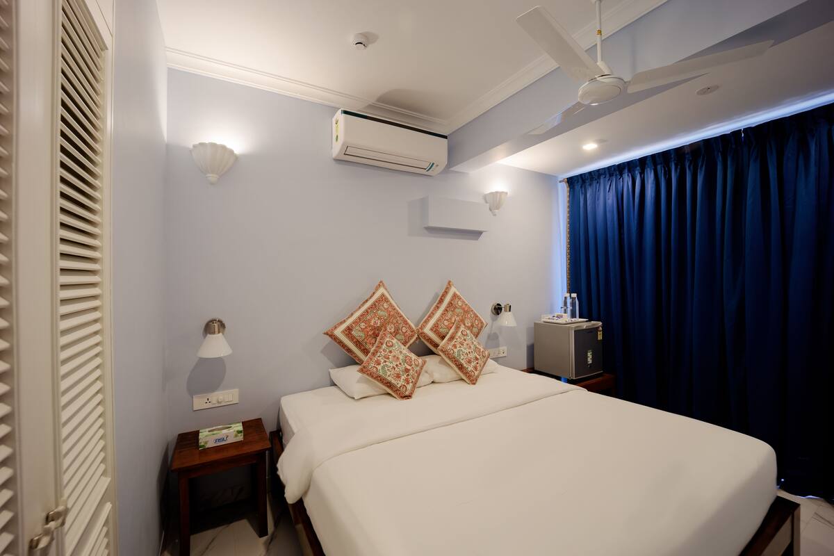 Quarto de hotel com cama, duas mesas de cabeceira, ar condicionado, ventoinha no teto, luminária na parede e janela com cortinas.