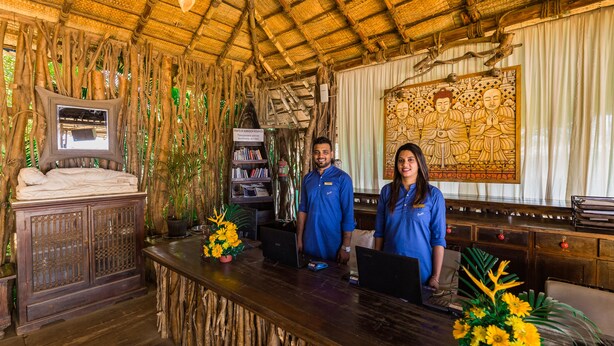 A reception area with a wooden desk, two staff members, a mirror, a bookshelf, and a Buddha painting. - Fiesta Beach Resort (Baga)
