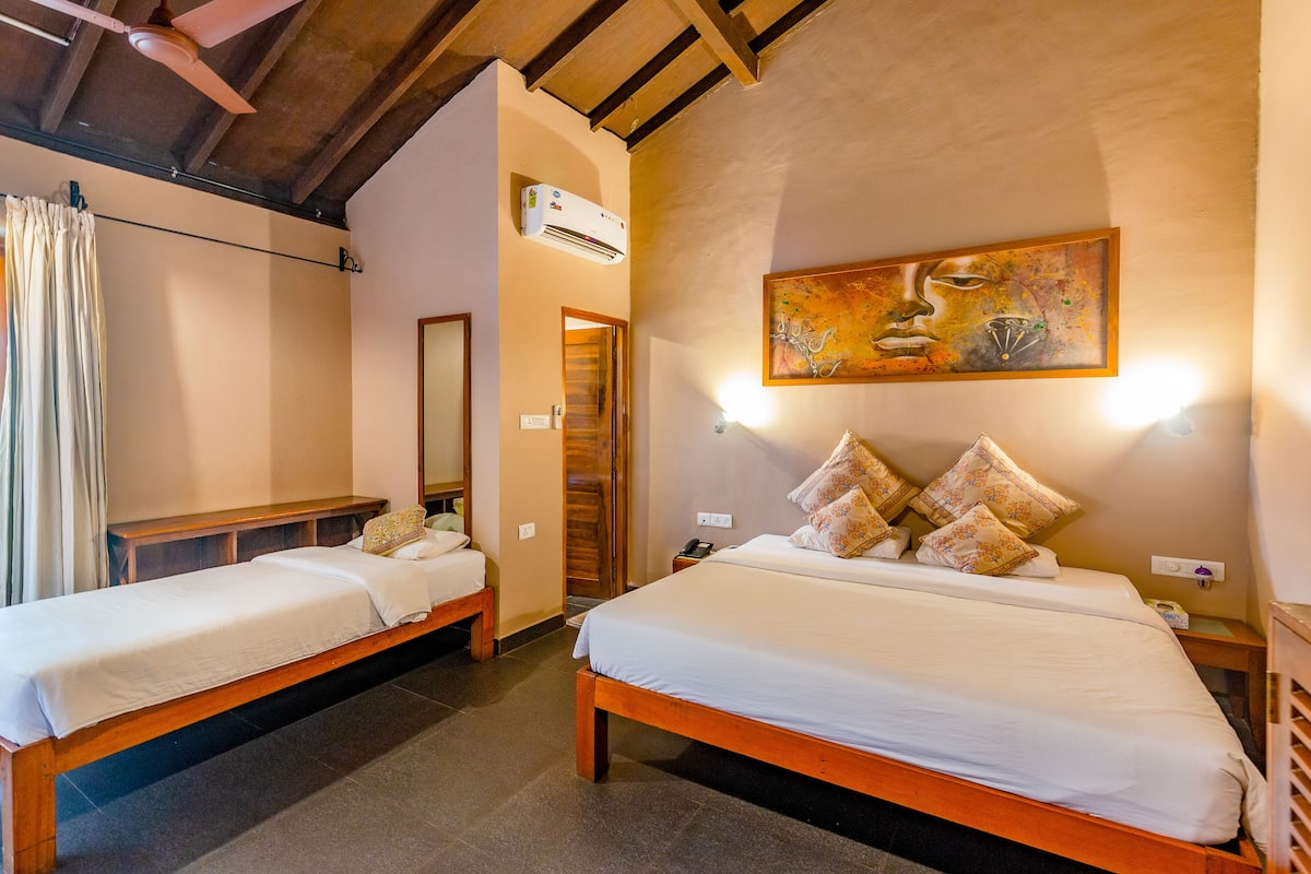 Quarto com duas camas de madeira, um ventilador de teto, um ar condicionado, um quadro e uma mesa de cabeceira com abajur.