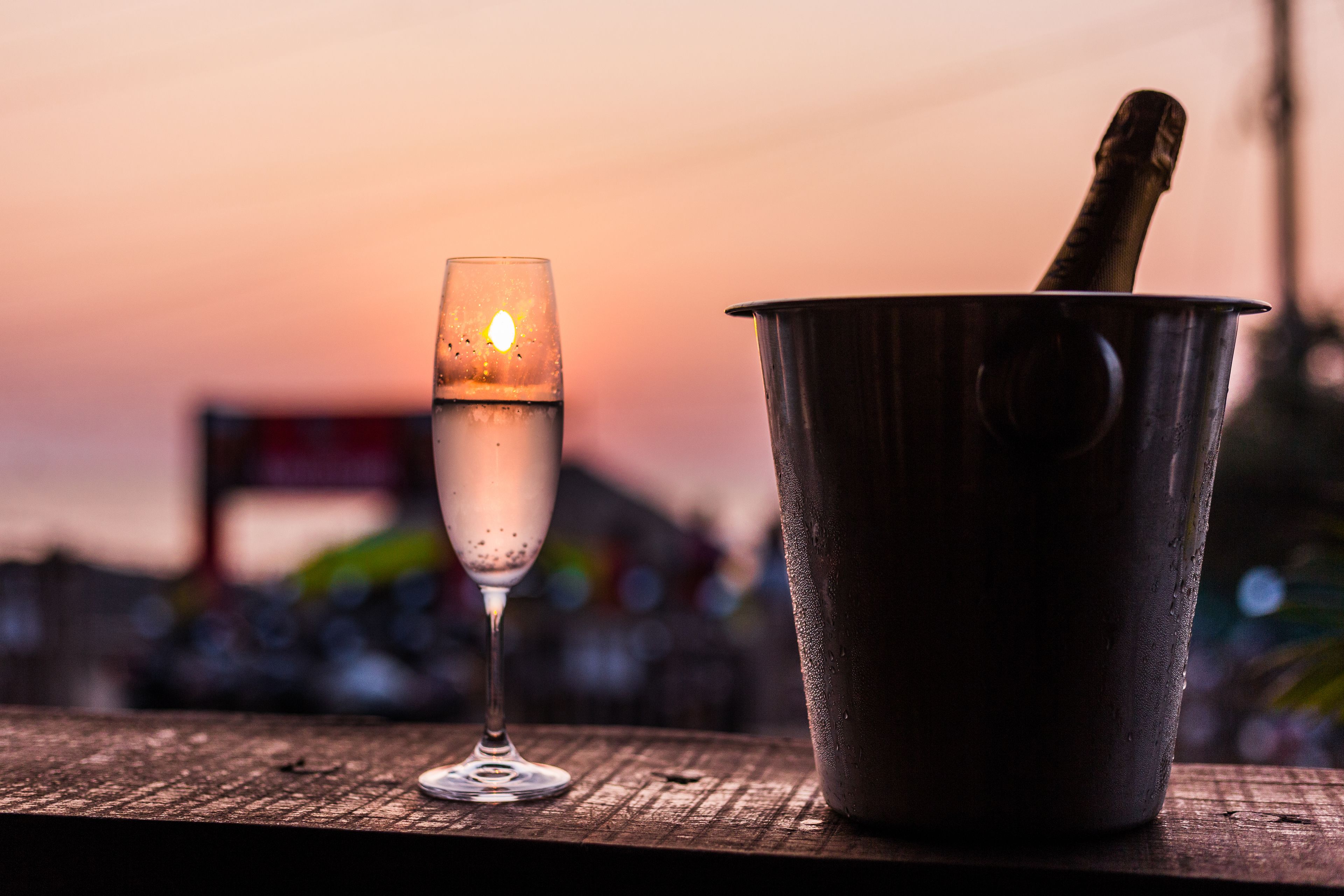 A glass of champagne and a champagne bucket on a wooden table with a sunset in the background.
