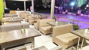 A restaurant with outdoor seating, including tables and cushioned benches, with a view of the street and parked cars. - Hotel Yog Palace (Rahata)