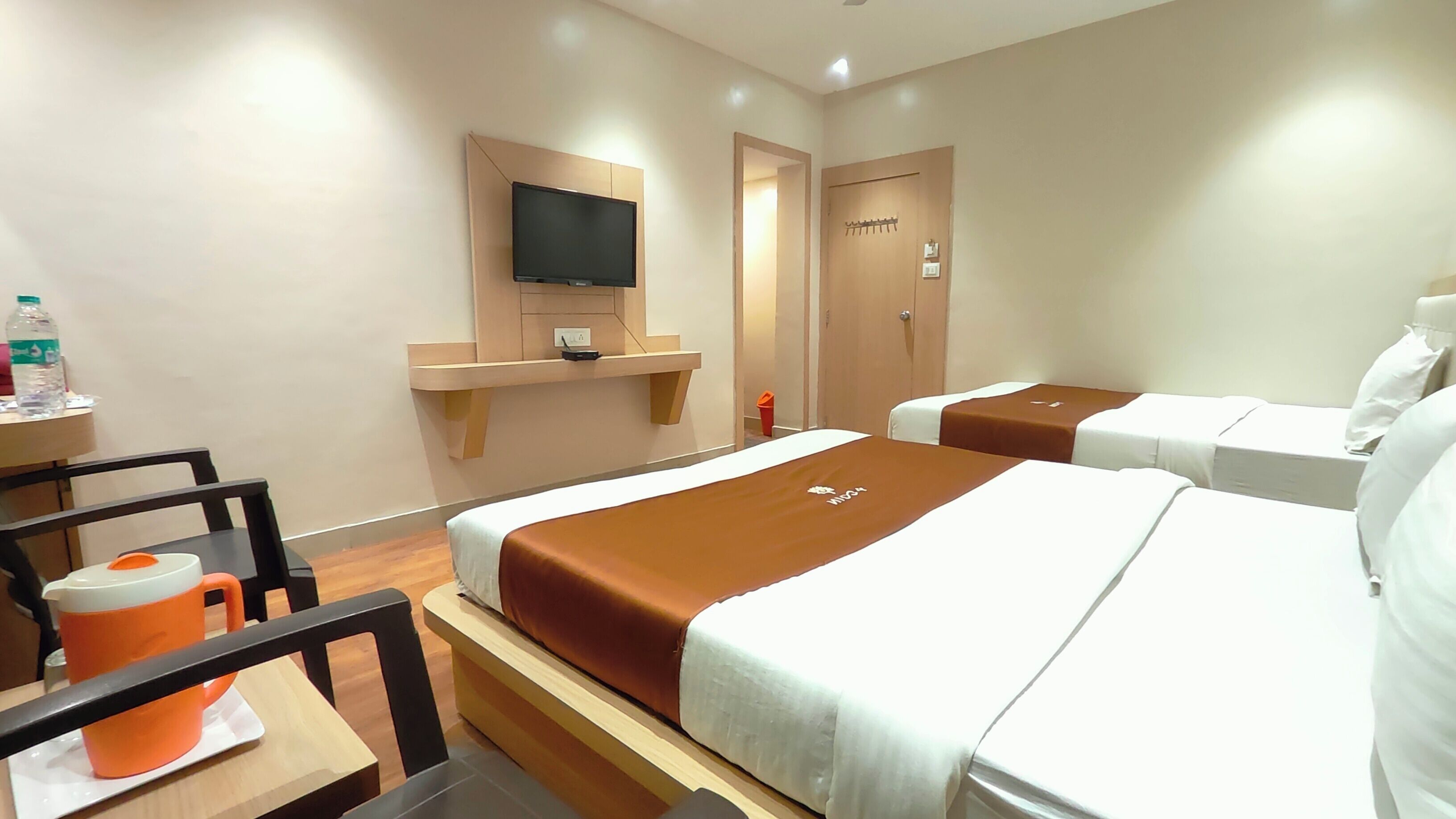 A hotel room with two beds, a TV mounted on the wall, a small table with a water bottle, and a chair.