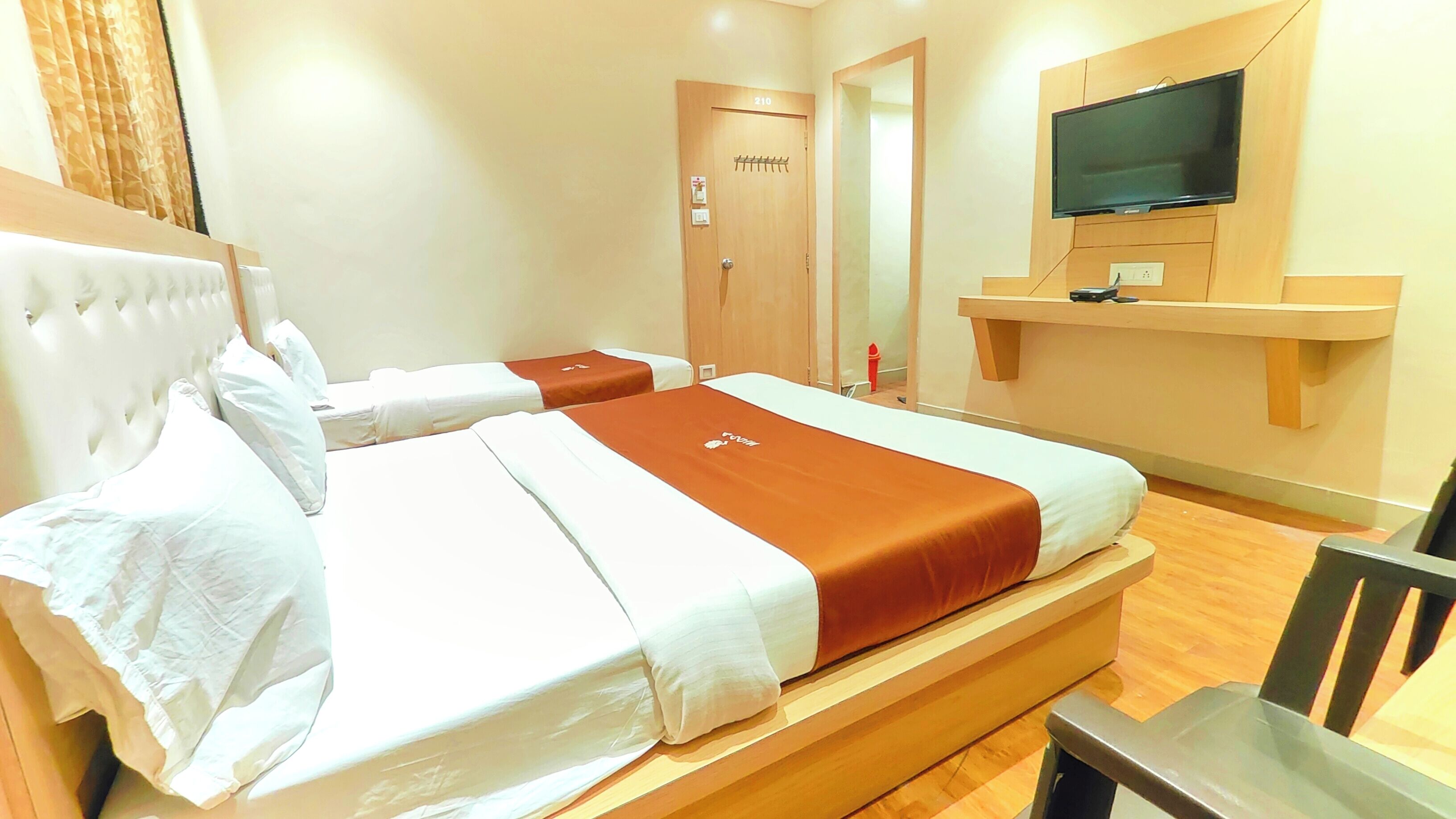 Ein Hotelzimmer mit zwei Betten, einem an einer Holzhilfe angebrachten Fernseher und einer Tür mit der Bezeichnung '210'.