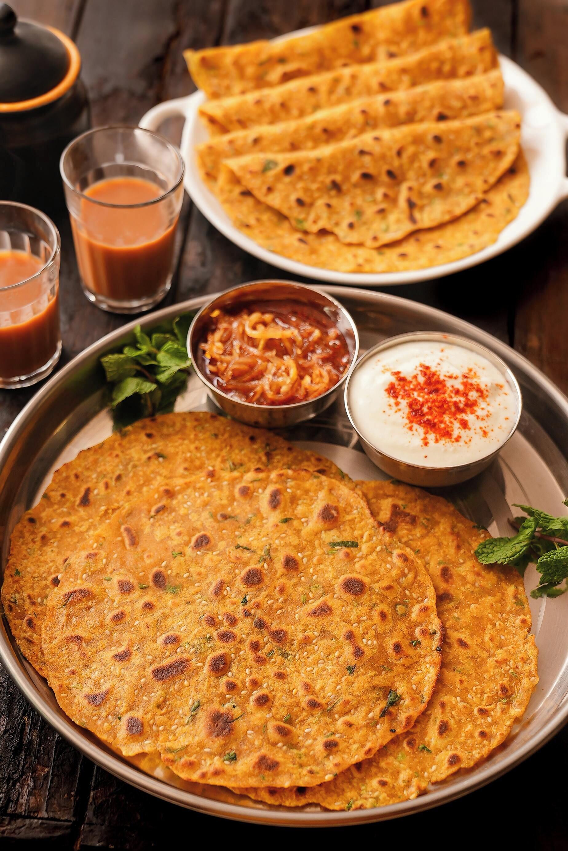 A plate of flatbreads with a side of chutney and yogurt, and two glasses of a brown beverage.