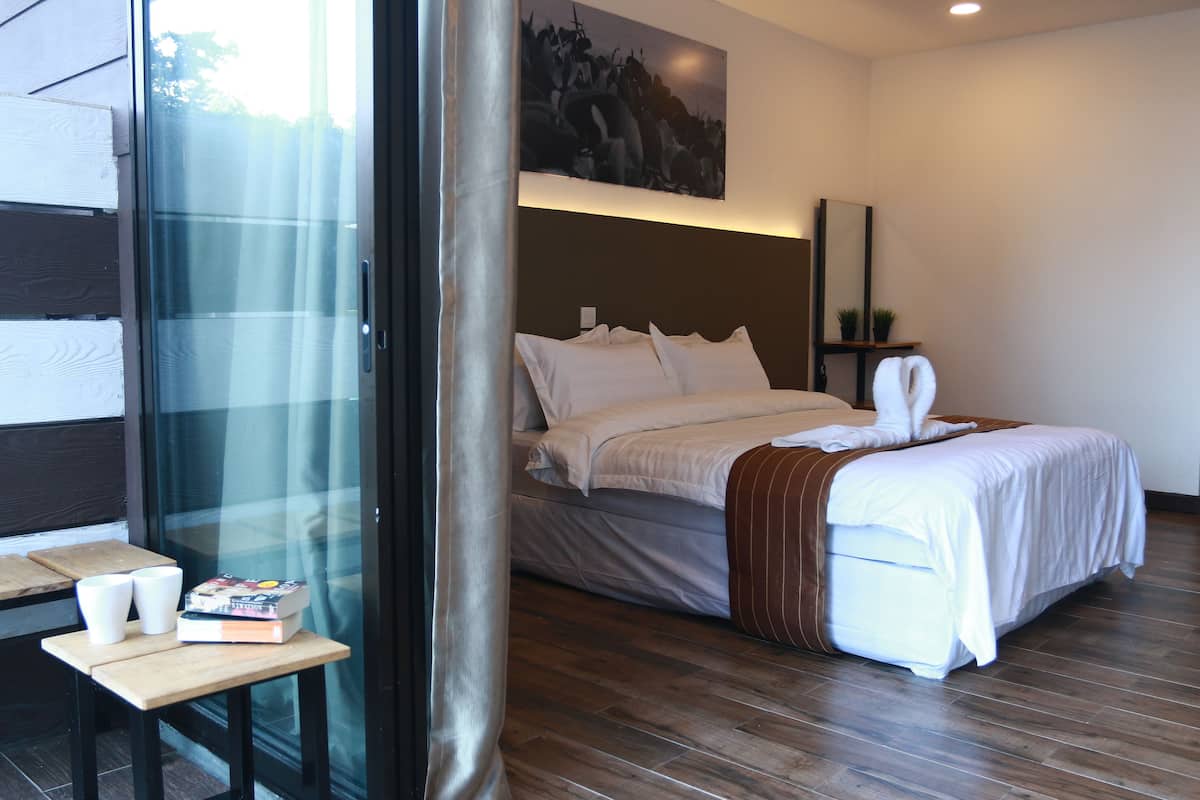 Habitación de hotel con una cama, una mesita de noche con un libro y gafas, una pequeña mesa con dos tazas y un cuadro en la pared.