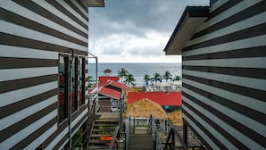 Vista desde un balcón de madera con vista a una playa, palmeras y un edificio de techo rojo.