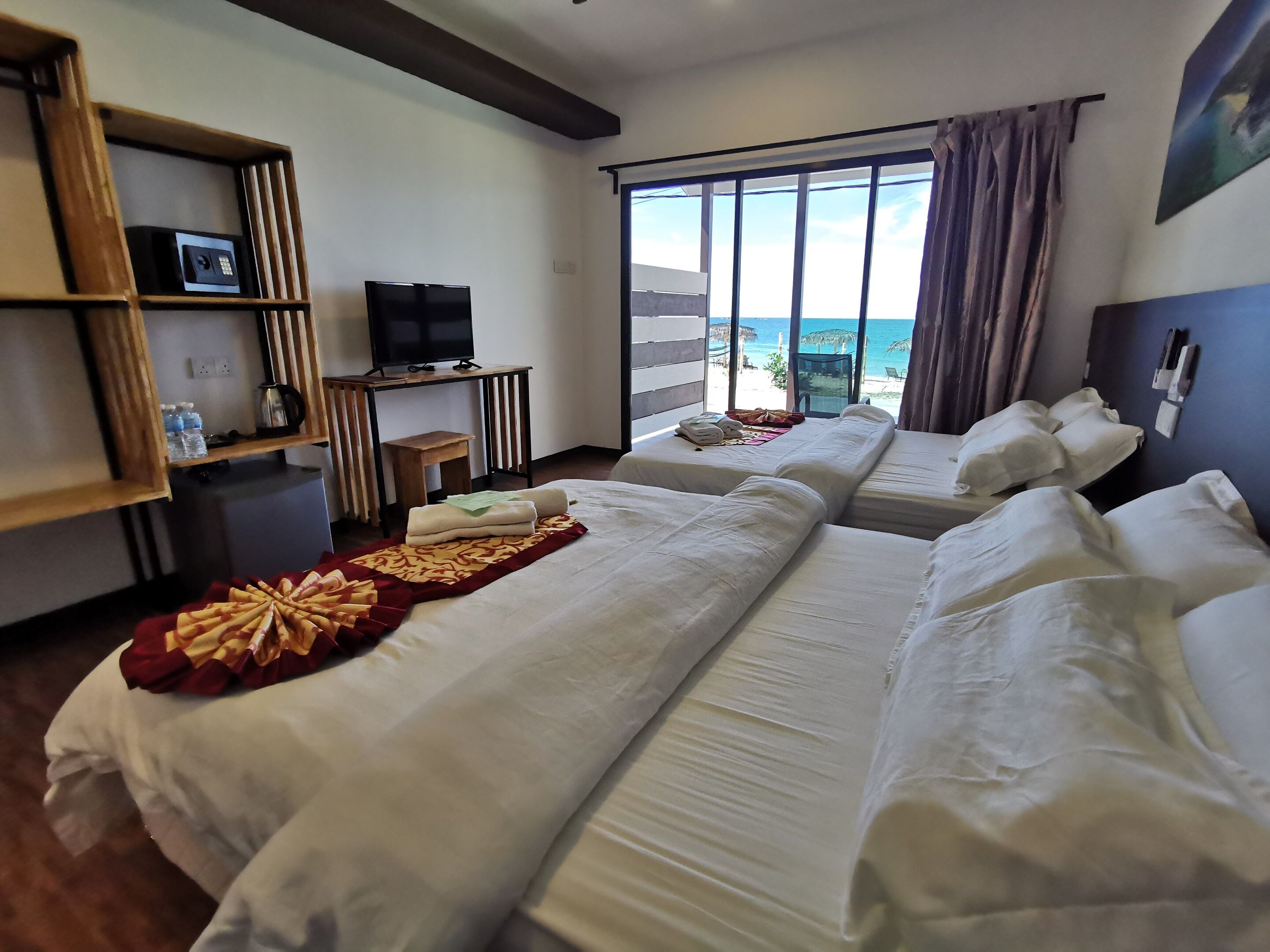 A hotel room with two beds, a TV, a desk, and a view of the beach.