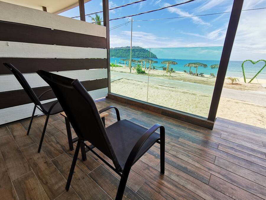 Una habitación con un gran ventanal con vista a la playa, suelo de madera y dos sillas negras.