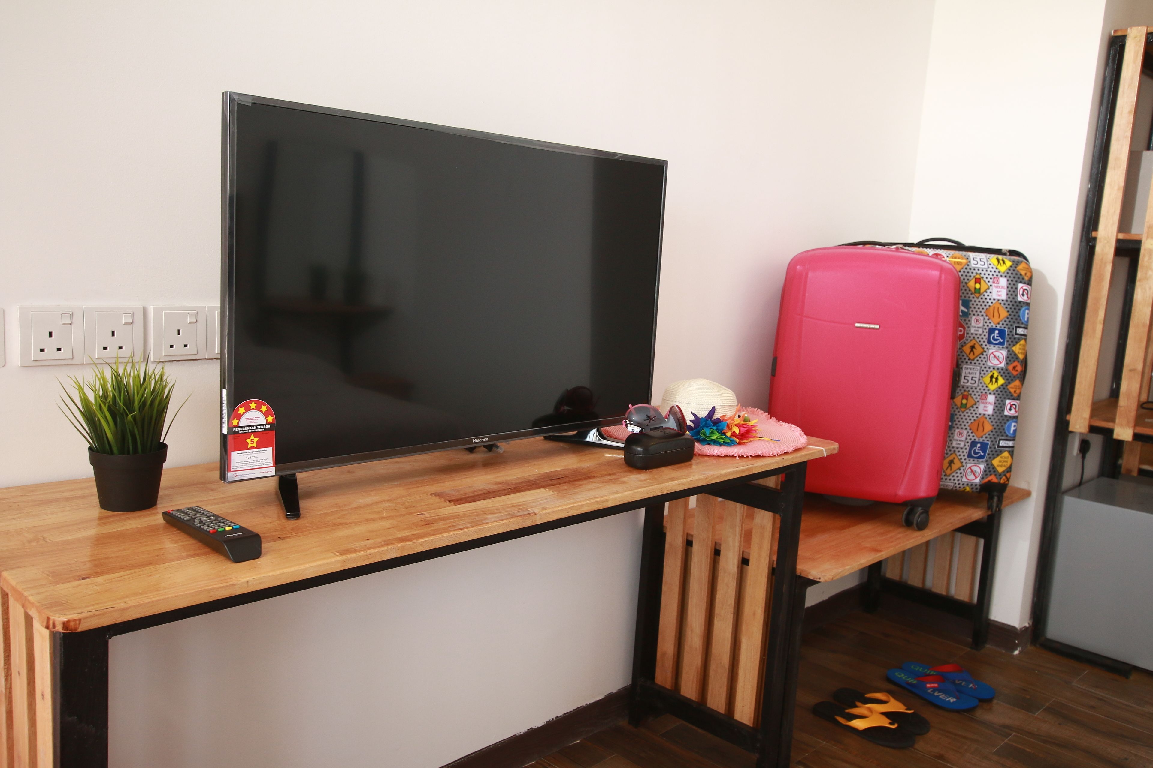 Een houten bureau met een flatscreen televisie, een afstandsbediening, een kleine potplant, een roze koffer en een koelkast.