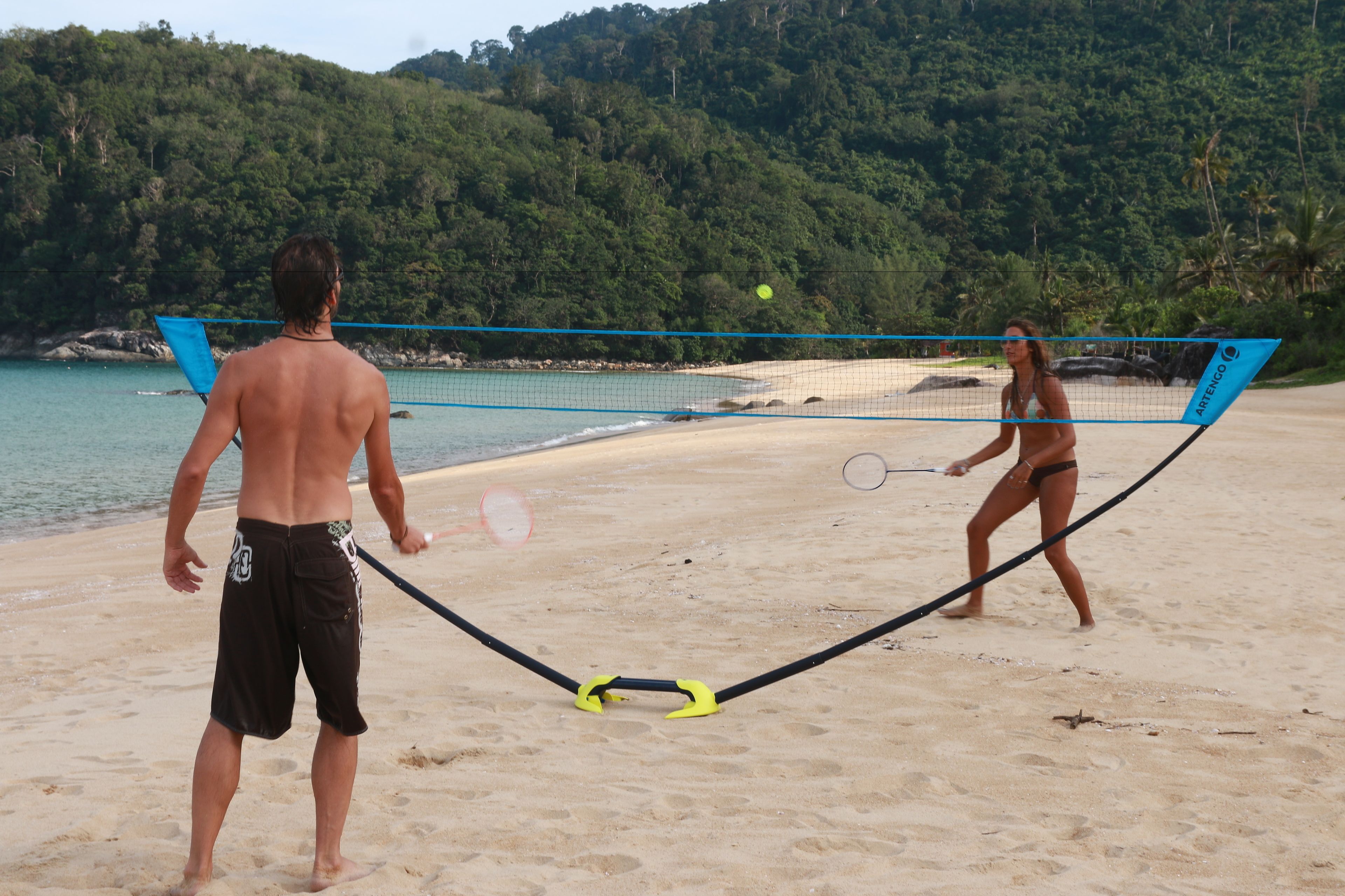 A beach volleyball game with a net and players on a sandy beach with a forested hill in the background.