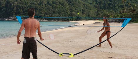 A beach volleyball game with a net and players on a sandy beach with a forested hill in the background.