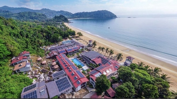 Vista aérea de un complejo turístico costero con varias edificaciones, una playa y una zona boscosa.