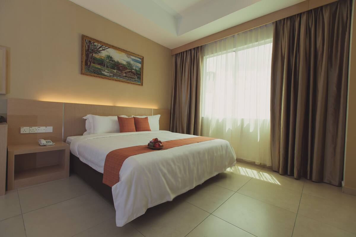 A hotel room with a large bed, a painting on the wall, a bedside table with a phone, and a window with curtains.