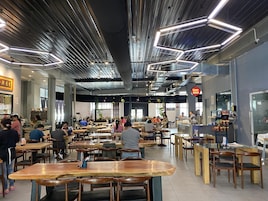 Um restaurante espaçoso com pé-direito alto, dutos aparentes e luminárias modernas. Mesas e cadeiras dispostas para refeições, e um balcão visível ao fundo.