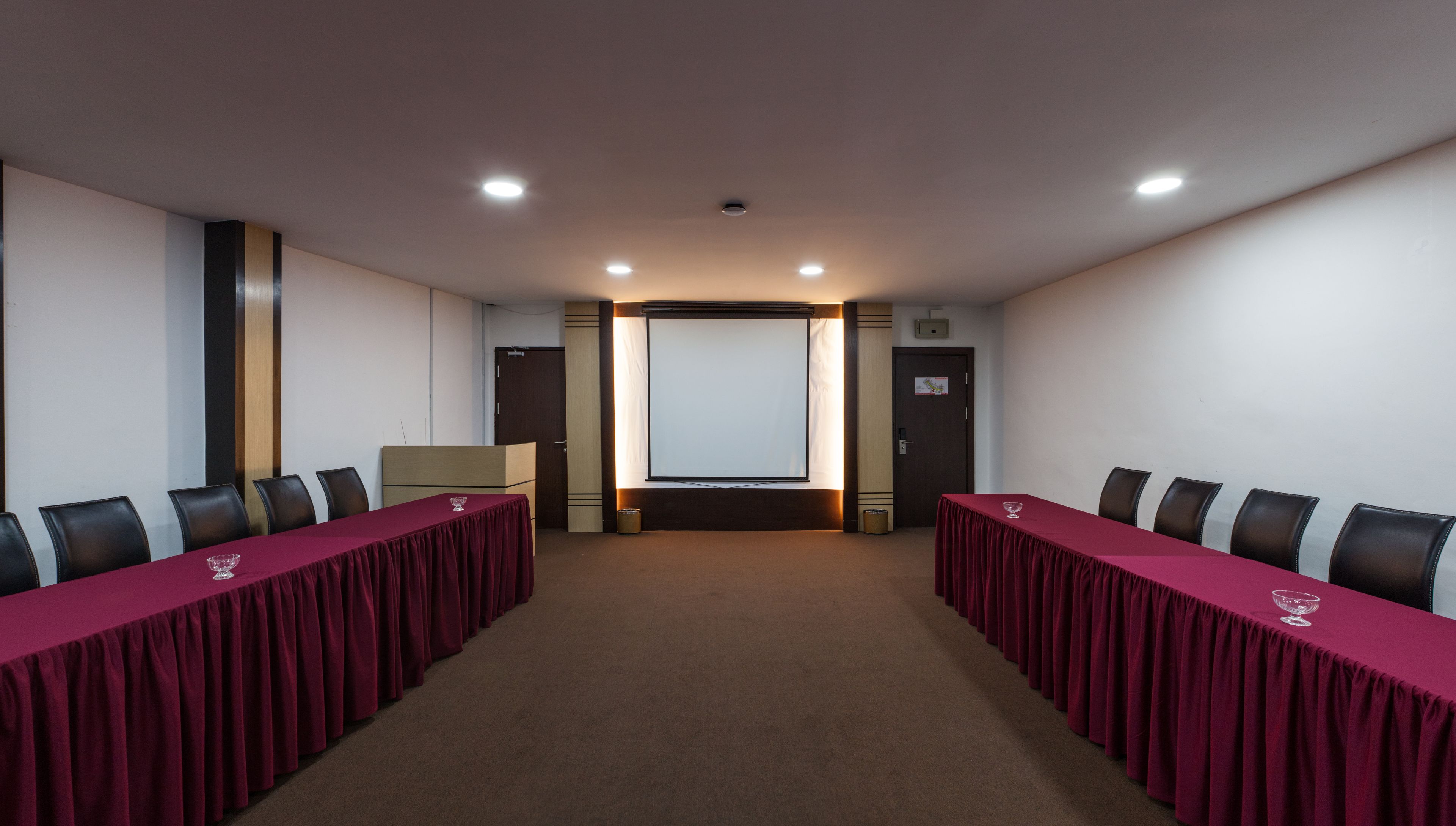 A conference room with two long tables, black chairs, a projector screen, and a door.