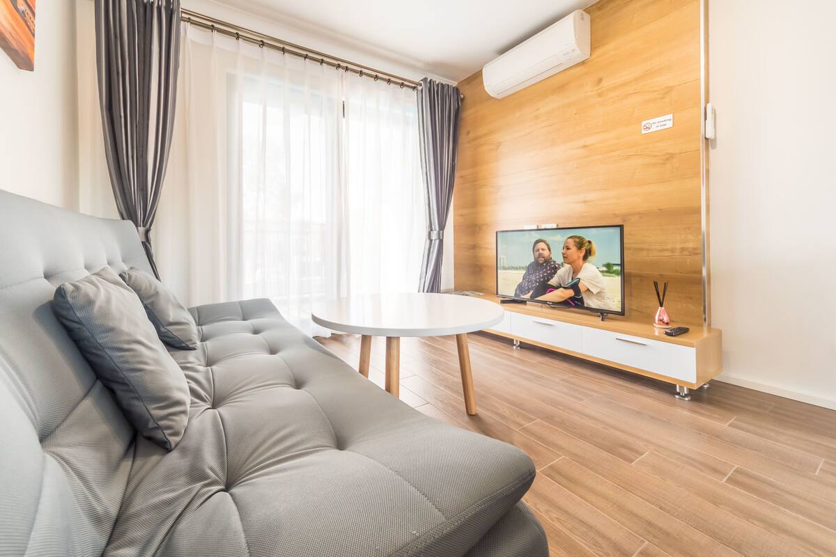 Uma sala de estar moderna com um sofá cinzento, uma mesinha branca e uma televisão em uma parede de madeira.