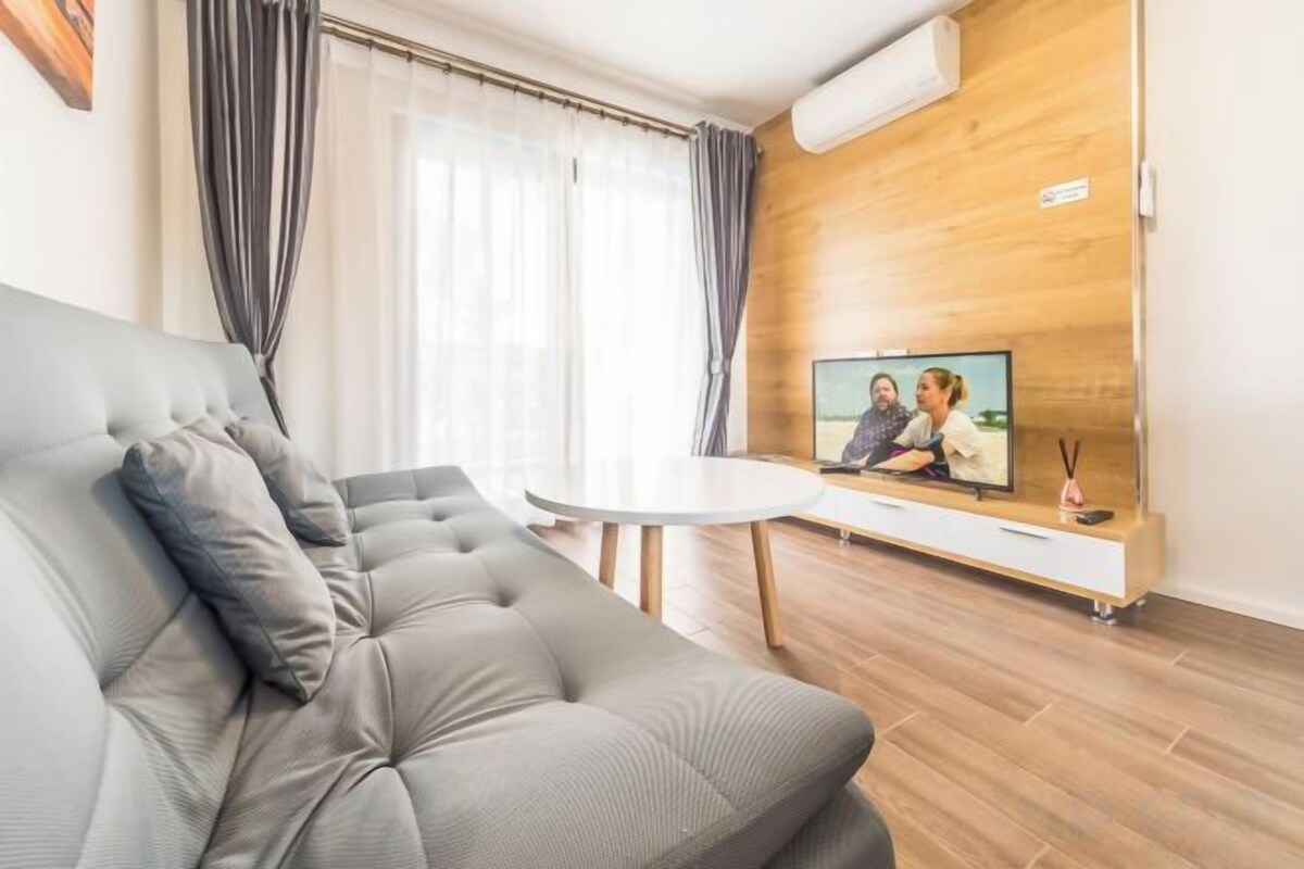 A modern living room with a grey sofa, a small round table, and a TV on a wooden wall.