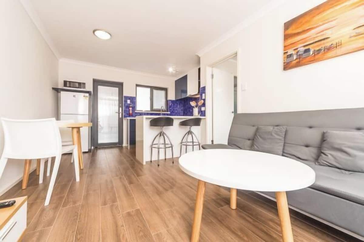 Sala de estar moderna com um sofá cinzento, uma mesa redonda branca e uma área de cozinha com frigorífico, fogão e banquetas.