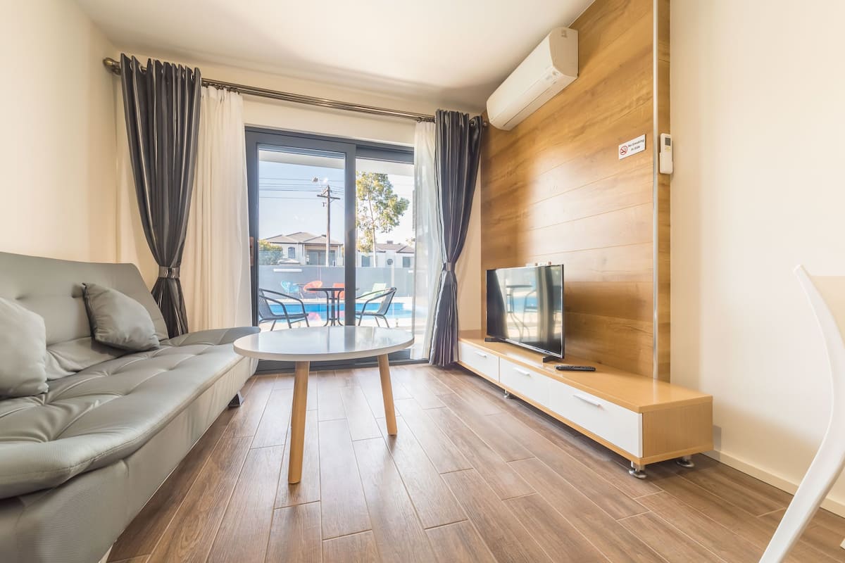 Uma sala de estar moderna com um sofá cinza, uma mesa redonda, uma TV em um armário de madeira, e vista para uma piscina através de uma porta de vidro.