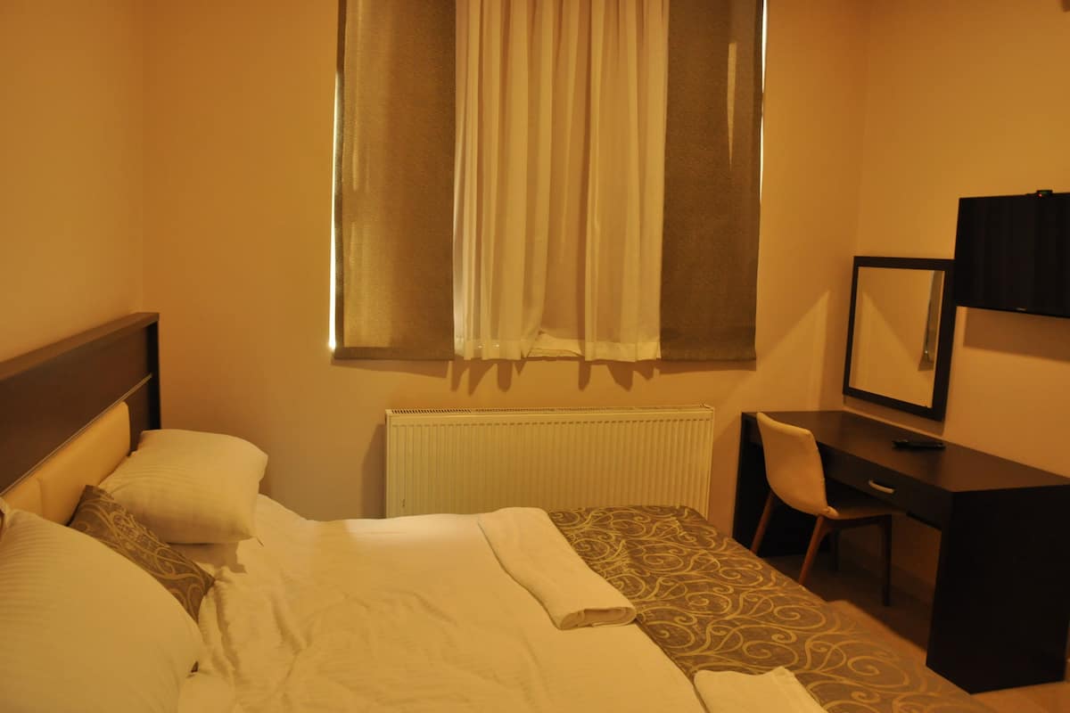 A hotel room with a bed, a desk, a chair, a window with curtains, and a radiator.