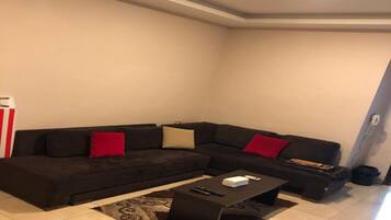 Een moderne woonkamer met een donkerbruine hoekbank, twee rode kussens, een zwarte salontafel en een kamerbreed tapijt met een patroon.