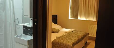 A bedroom with a bed, a nightstand, a window with curtains, a radiator, and a bathroom with a shower and sink.