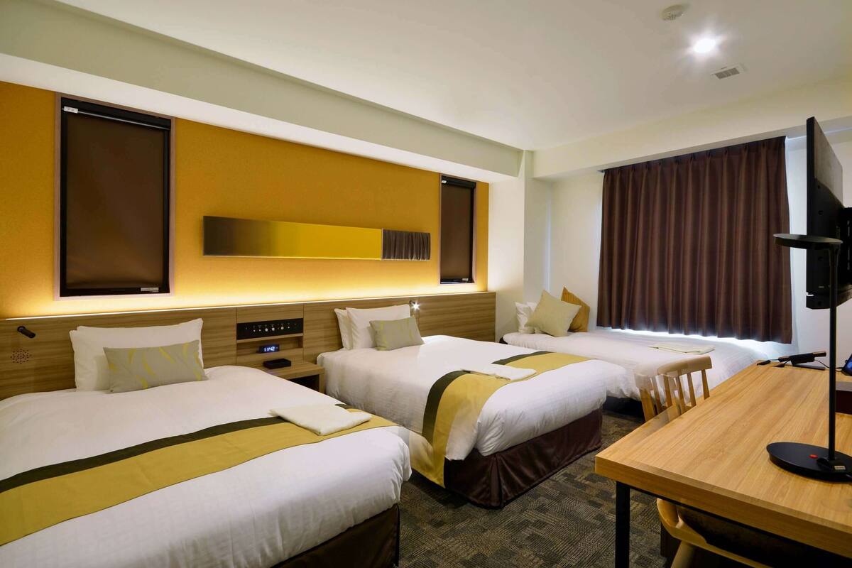 Ein Hotelzimmer mit drei Betten, einem Holztisch und einem großen Spiegel an der Wand.