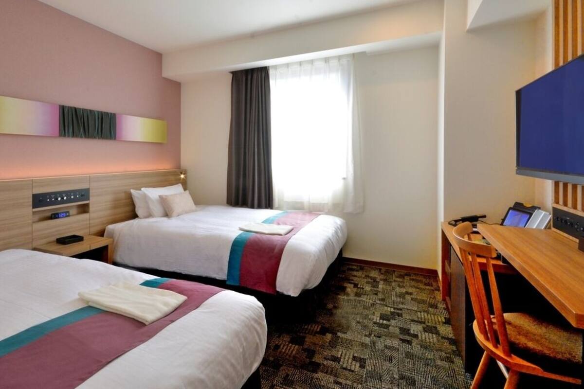 Ein Hotelzimmer mit zwei Betten, einem Schreibtisch, einem Stuhl, einem Fernseher und einem Fenster mit Vorhängen.
