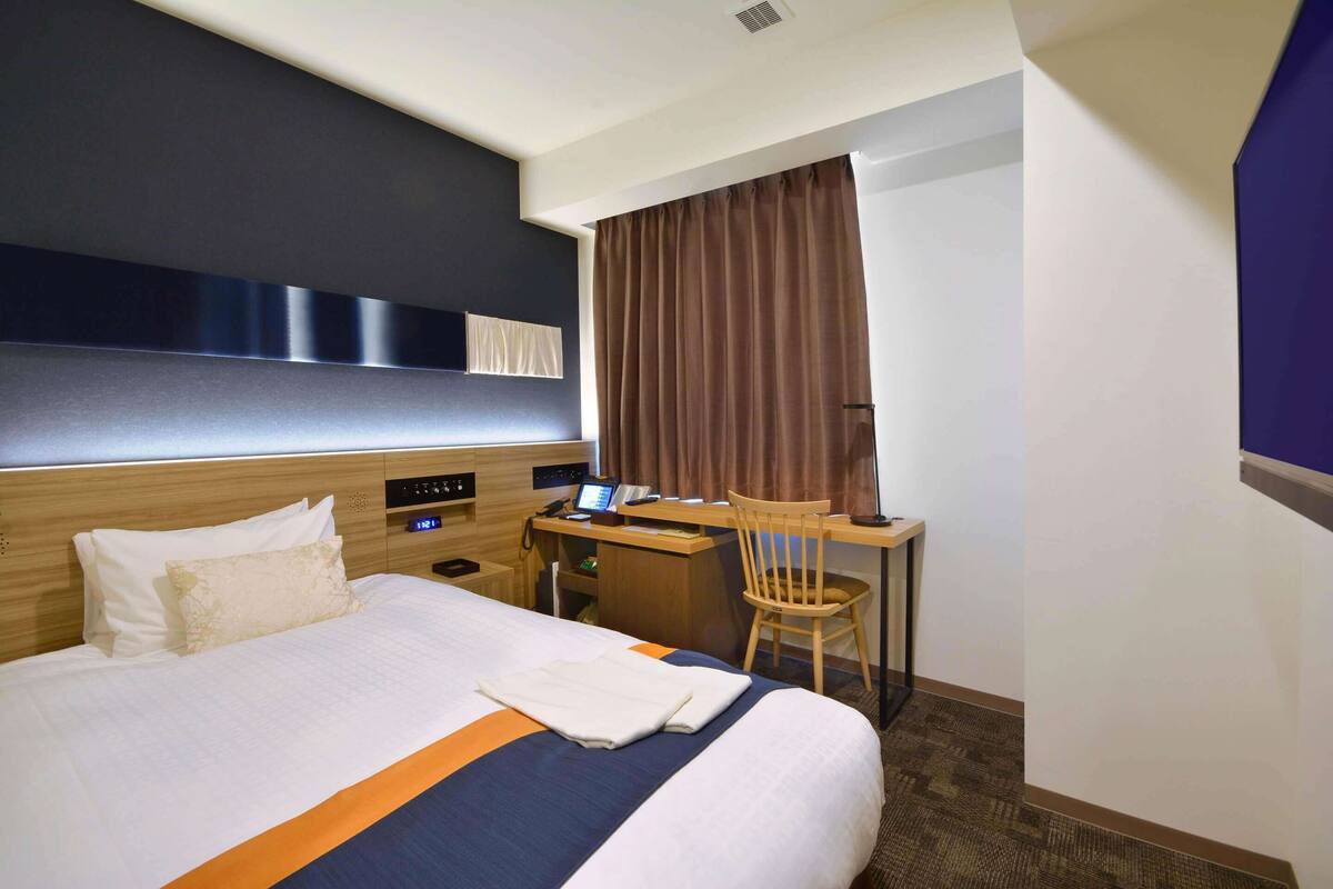 Ein Hotelzimmer mit einem ordentlich bezogenen Bett, einem Schreibtisch mit Stuhl und einem großen Fenster mit Vorhängen.