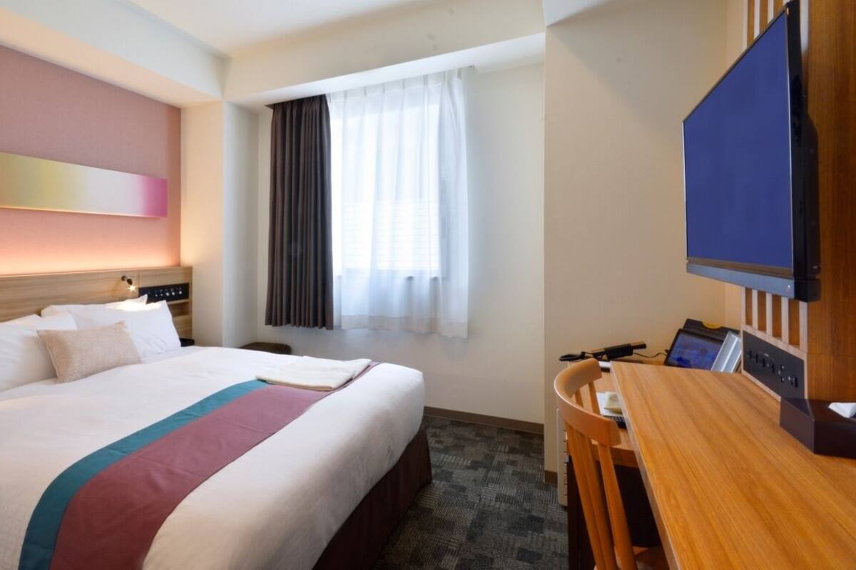 Ein Hotelzimmer mit Bett, Schreibtisch, Fernseher und einem fensterbepflanzten Fenster.