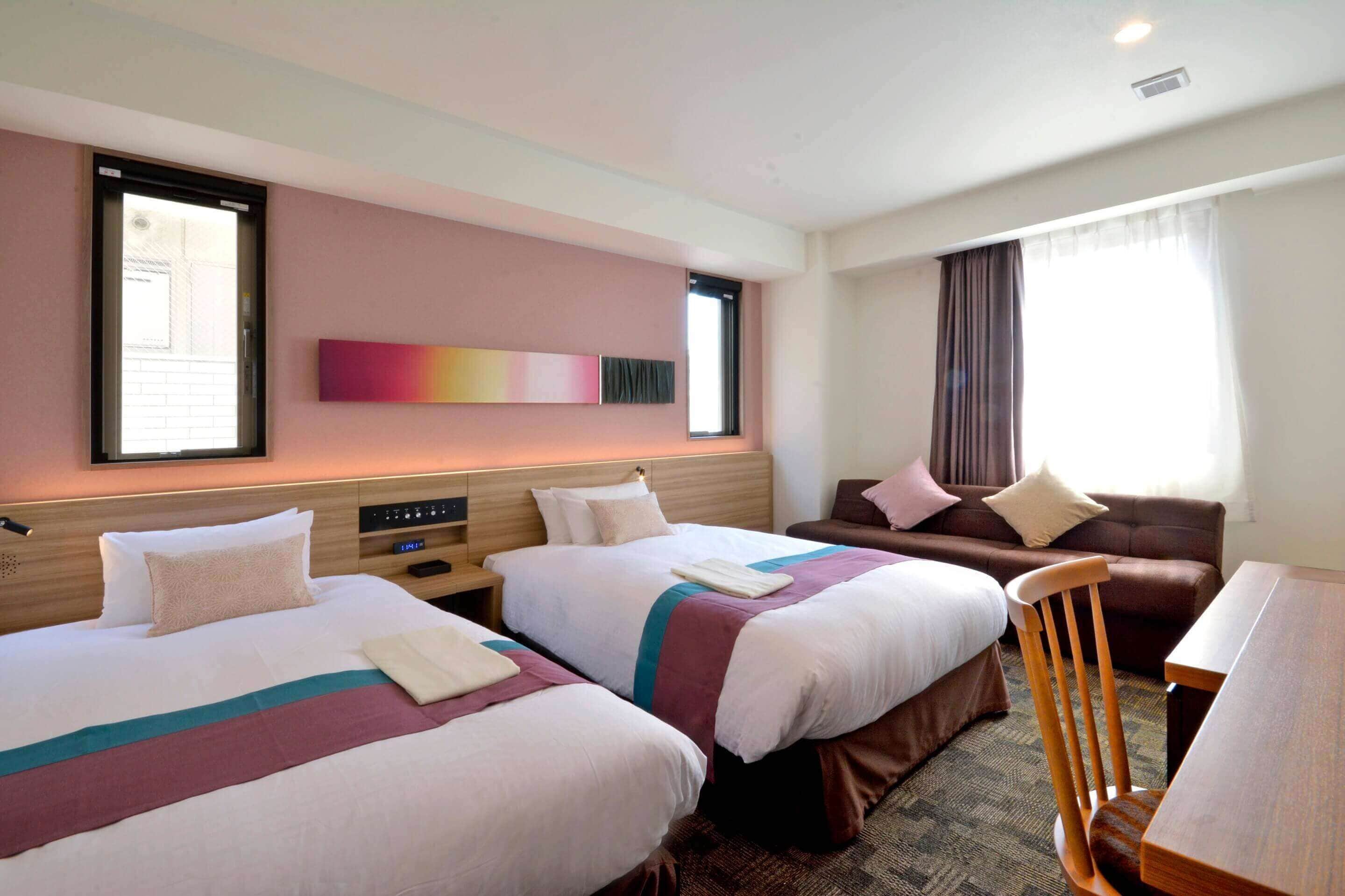A hotel room with two beds, a sofa, a wooden chair, a desk, and a large window with curtains.