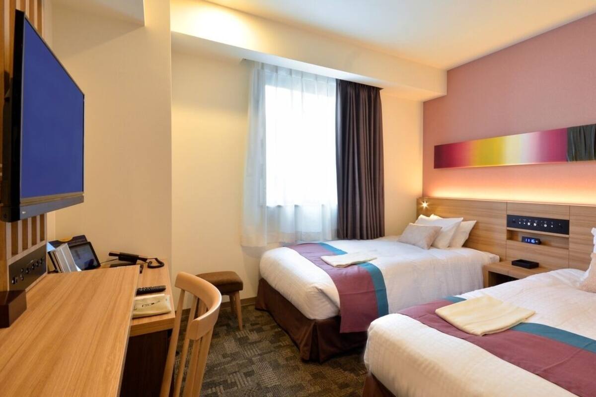 Ein Hotelzimmer mit zwei Betten, einem Schreibtisch, einem Fernseher und einem Fenster mit Vorhängen.