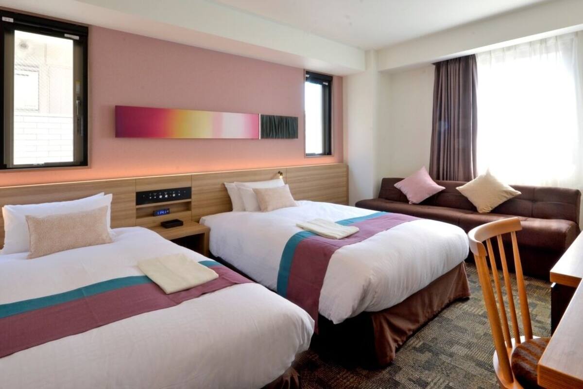 Une chambre d’hôtel avec deux lits, un canapé, une chaise en bois, un bureau et une fenêtre avec des rideaux.
